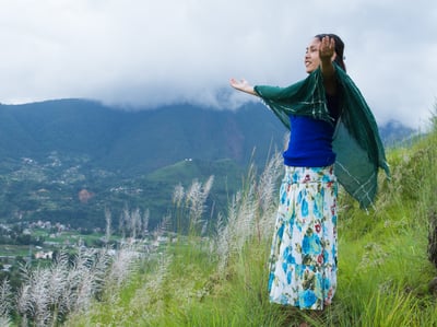 Girl extends hands with a smile, green scarf billowing in the wind, standing on a lush green hillside