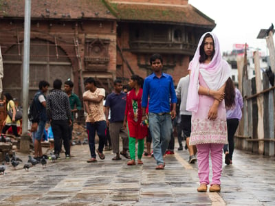 Girl in pink traditional Asian clothes and head scarf stands in a busy area waiting for someone.