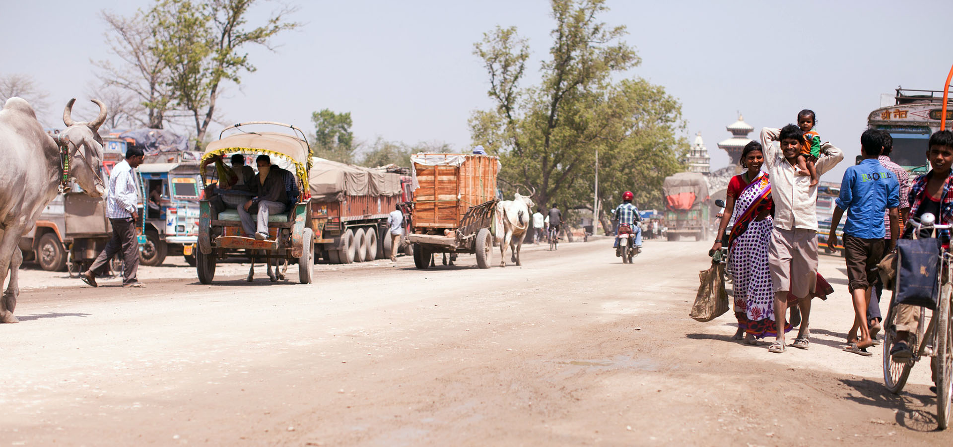 People walking down a dirt road, smiling; other vehicles and animals move down the road as well.